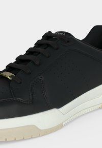 Baskets en cuir noir avec lacets noirs, détails perforés sur le côté, semelle en caoutchouc blanche et petite étiquette dorée sur les lacets sur un fond uni.