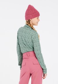 Camicia verde con pois neri, maniche lunghe e colletto, abbinata a pantaloni rosa a vita alta e a un berretto lavorato a maglia rosa testurizzato.