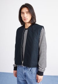 Veste sans manches bleu marine foncé avec fermeture éclair à l'avant, grandes poches et col côtelé, portée par-dessus une chemise à manches longues rayée noir et blanc.