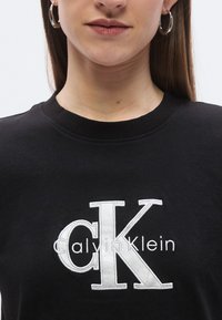 Svart bomullsgenser med rund hals, med stor hvit "CK"-logo og "Calvin Klein"-tekst brodert på forsiden.