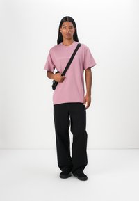 Lichtroze t-shirt met korte mouwen en een eenvoudige witte graphic, gecombineerd met losse zwarte broek en zwarte schoenen, en een zwarte schoudertas.