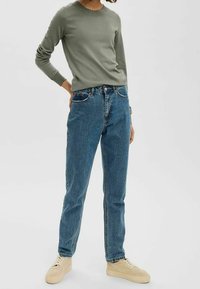 Pull vert olive à manches longues avec encolure festonnée, associé à un jean bleu taille haute et des baskets beige. Tenue simple et décontractée.