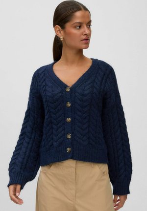 Femme portant un cardigan bleu marine en maille torsadée boutonné, manches longues et pantalon beige, regardant sur le côté sur un fond uni.