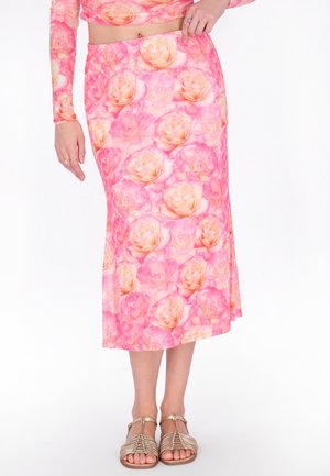 Vrouw draagt een roze midi-rok met bloemenprint en een bijpassende top met lange mouwen, met gouden platte sandalen met bandjes, op een witte achtergrond.