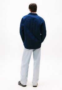 Vyras stovi su rankomis kišenėse, vilkėdamas tamsiai mėlyną dvigubą striukę, šviesiai mėlynus džinsus ir juodus batus, atsisukęs į šalin ant balto fono.