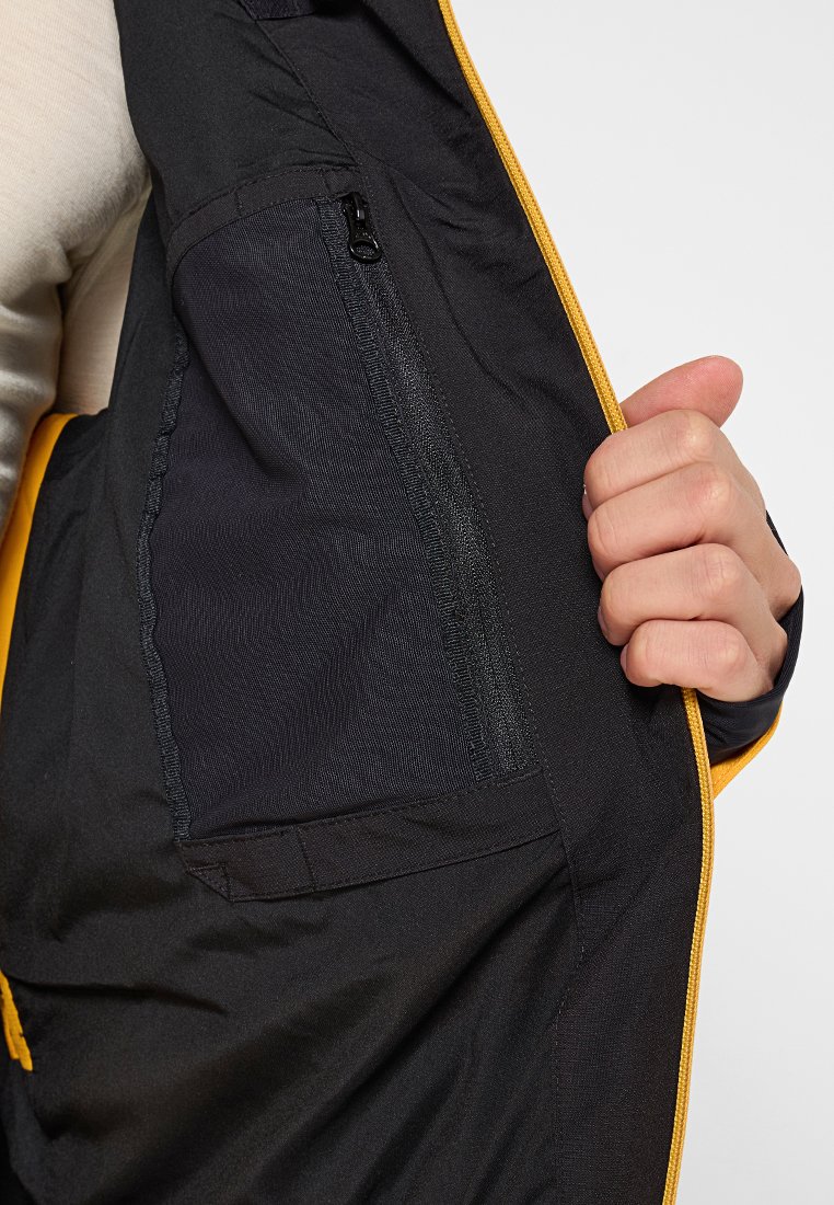 Giacca nera con zip e dettagli gialli, che mostra un tessuto testurizzato e una tasca con zip laterale. Presenta cuciture dettagliate e una vestibilità rilassata.