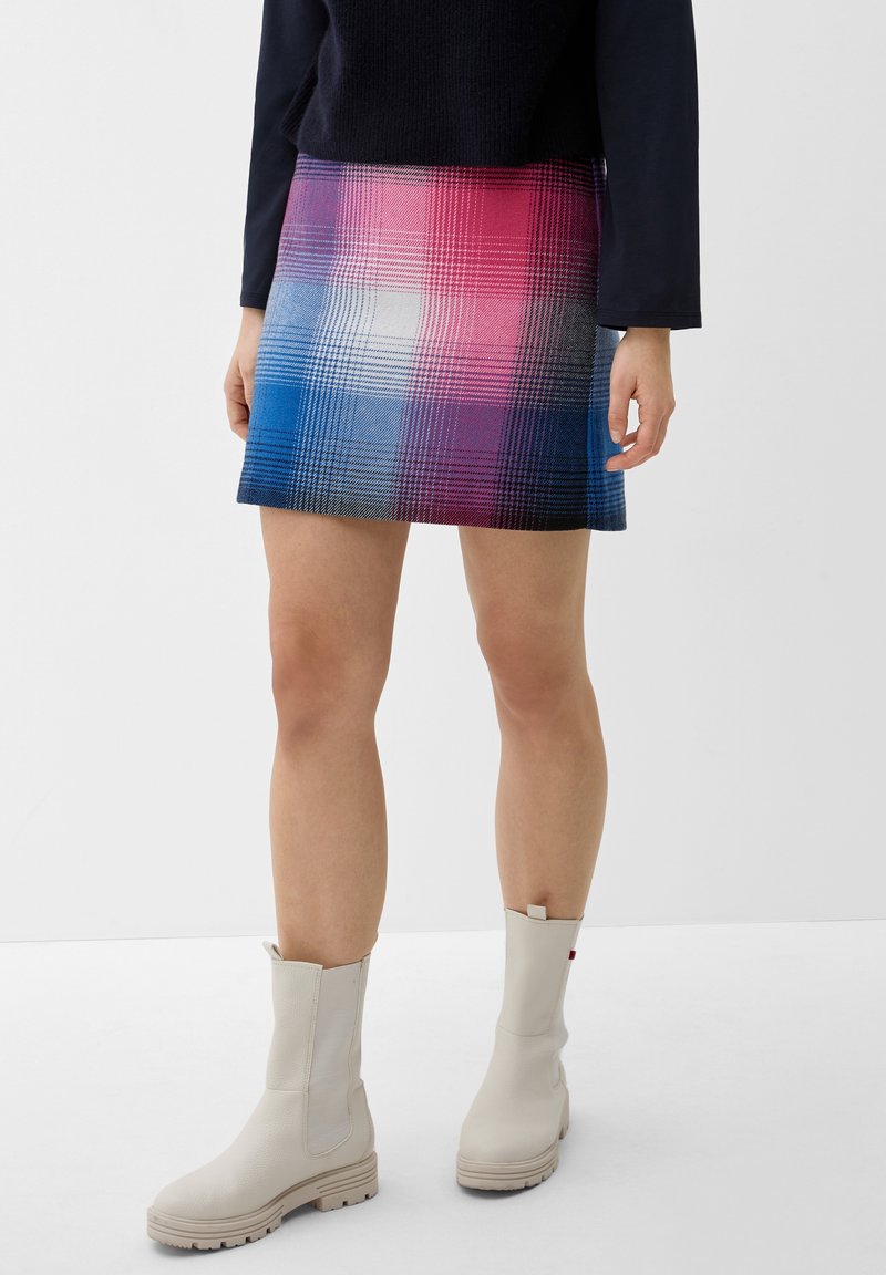Femme portant une mini-jupe à carreaux multicolores rose et bleu, des bottes blanches montant jusqu'à mi-mollet et un haut sombre à manches longues, debout sur un sol blanc.