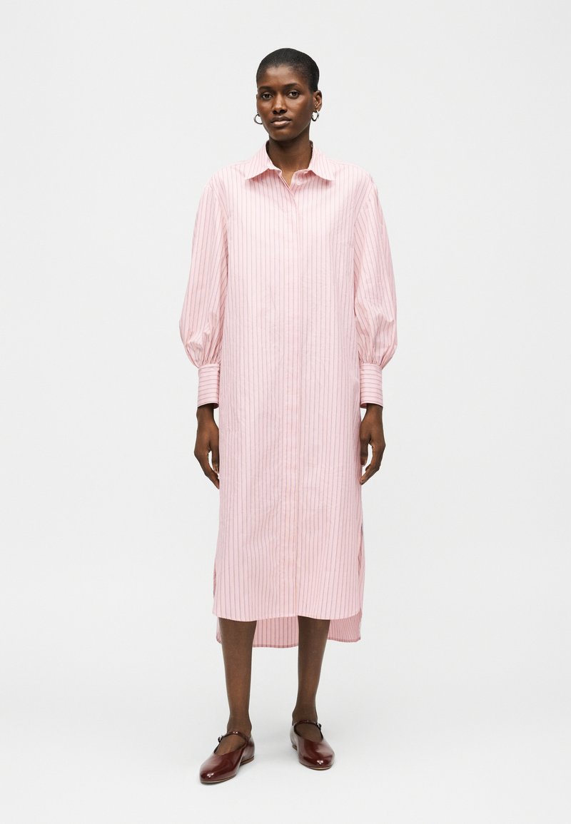 Γυναίκα που φοράει μακρύ ρόζ ριγέ πουκάμισο-φόρεμα με φουσκωτά μανίκια, μανσέτες με κουμπιά και καφέ επίπεδα παπούτσια, στέκεται μπροστά σε απλό λευκό φόντο.