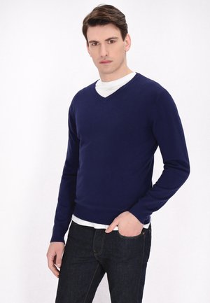Navyblauwe v-hals trui van zacht materiaal, met lange mouwen en een slim fit, gedragen over een wit shirt en gecombineerd met donkere jeans.