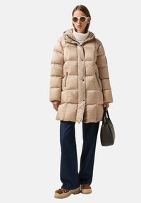 Beige Steppjacke mit Kapuze, Reißverschlusstaschen und Knopfverschluss. Kombiniert mit dunkelblauen Schlagjeans und hellbraunen Sneakers.