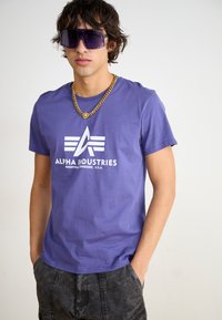 Violeta kokvilnas T-krekls ar baltu logotipu un tekstu "ALPHA INDUSTRIES" priekšpusē, saskaņots ar masīvu zelta ķēdes kaklarotu un lielām saulesbrillēm.