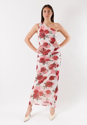 Donna in piedi con le mani sui fianchi, che indossa un vestito bianco monospalla con stampa floreale rossa e rosa e tacchi alti beige.