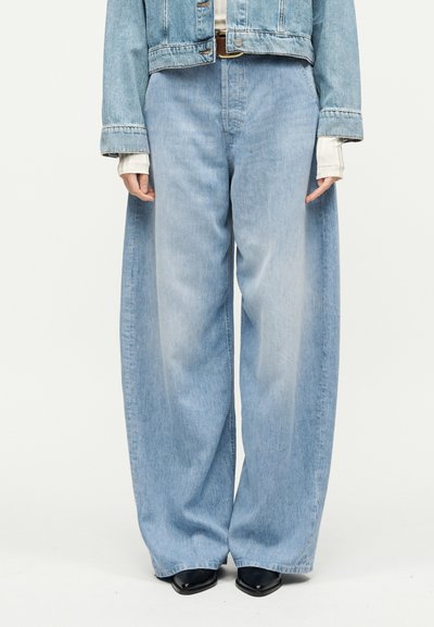 Άτομο που φοράει φαρδιά τζιν παντελόνια σε ανοιχτό μπλε χρώμα, κοντό τζιν μπουφάν, λευκό πουκάμισο με μακριά μανίκια από μέσα και μαύρα παπούτσια με μυτερή μύτη, όρθιο.