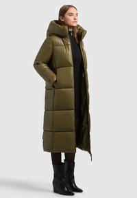 Lange olijfgroene pufferjas met een gequilte design, oversized capuchon, ritssluiting en zijzakken; gedragen over een zwarte jurk en gecombineerd met zwarte laarzen.