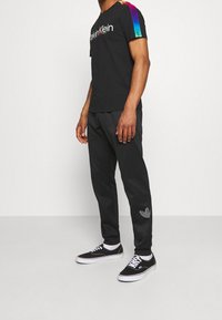 Černé tričko s barevným logem Calvin Klein na rameni a černé tepláky s malým vyšitým logem Adidas. Černé tenisky.
