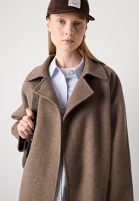 Ung kvinde iført en stor, brun uldfrakke, lyseblå skjorte med knapper, mørkebrun kasket, gyldne hoops og bærende en mørk skuldertaske.
