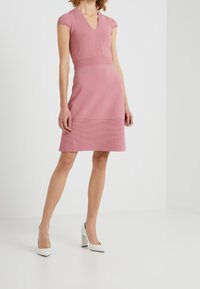 Rosa gestricktes Kleid mit V-Ausschnitt und kurzen Ärmeln, das horizontale Rippungen an der Taille und dem Saum aufweist, kombiniert mit weißen High Heels.