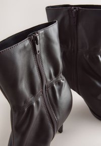 Stivaletti alla caviglia in pelle marrone scuro con zip laterali e dettagli leggermente plissettati, mostrati su uno sfondo chiaro.