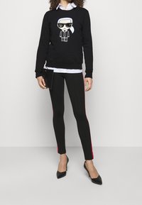 Sudadera negra con gráfica de dibujos animados, camisa blanca de botones debajo, leggins negros con tiras laterales rojas y tacones negros de punta.