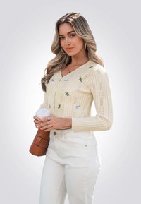 Crèmekleurige gebreide cardigan met bloemembroderie, lange mouwen en een V-hals. Gepaard met witte jeans en een koffiekopje vast.