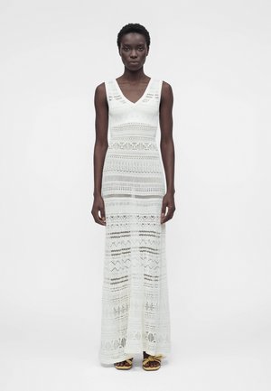 Slank sort kvinde iført en ærmeløs, hvid hæklet maxi-kjole med V-hals og knyttede sandaler, stående mod en ensartet lys baggrund.