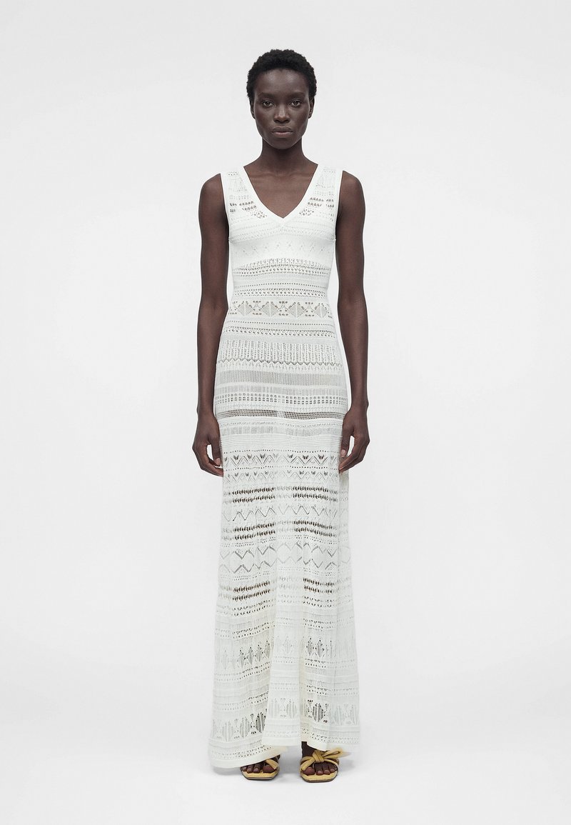 Λεπτή μαύρη γυναίκα που φοράει λευκό, αμάνικο πλεκτό φόρεμα μάξι με V-λαιμόκοψη και σανδάλια με κόμπους, στέκεται σε απλό ανοιχτό φόντο.