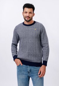Pulover u mornarsko plavoj i bijeloj boji s uzorkom riblje kosti, s rebrastim završecima na rukavima i donjem dijelu. Ima mali žuti logo na prsima. Nositi s plavim trapericama.