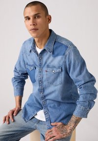 Camisa de denim azul claro con cuello en punta, dos bolsillos en el pecho y cierre con botones. Presenta una textura ligeramente deslavada y mangas largas.