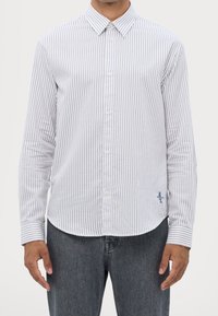 Camisa blanca de botones con rayas verticales azules, de mangas largas y cuello clásico. Presenta un pequeño logo en el dobladillo izquierdo.