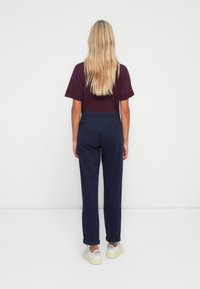Femme aux longs cheveux blonds, debout face à un mur blanc, vêtue d'une chemise violet foncé, d'un pantalon bleu marine et de baskets blanches avec le logo "VEJA".