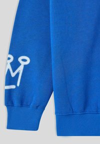 Niebieski rękaw sweatra z ribbowym mankietem, z białym logo korony na boku. Gładka tekstura materiału bez widocznych wzorów.