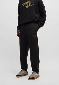 Sort bomulds hoodie med guldlogo, matchet med sorte sweatpants med et guldemblem. Grå sneakers med sorte og gummisåler.