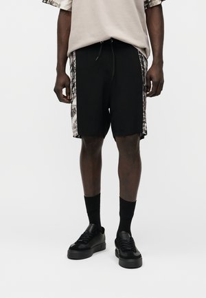 Homme portant un short noir avec des panneaux latéraux à motifs, des chaussettes noires et des baskets noires, debout devant un fond clair uni.