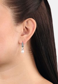 Kvinnas öra med silverörhänge som har en liten rund kristall och en dropformad vit pärla som hänger nedanför.