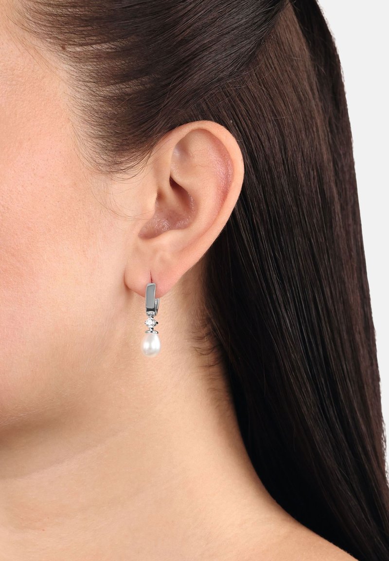 Kvinnas öra med silverörhänge som har en liten rund kristall och en dropformad vit pärla som hänger nedanför.