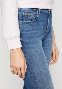Jeans de corte slim en denim de color azul, con costuras en contraste, diseño de cinco bolsillos y un ligero desgaste, combinados con un sudadera de color rosa claro.