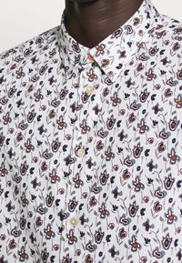 Bílá košile s knoflíky, která má květinový vzor v modré, červené a šedé barvě. Jemný materiál s klasickým límcem a perlovými knoflíky.