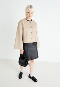 Giacca beige cropped con grandi bottoni dorati, abbinata a una mini gonna in pelle nera, mocassini in pelle verniciata nera e una borsa nera. Capelli corti.