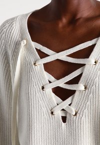 Crème kleurige gebreide trui met een diepe V-hals, gekruiste veteringang en goudkleurige ringen, zachte textuur en geribbelde details.