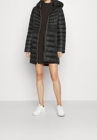 Schwarzer gesteppter Mantel mit Kapuze, ausgestattet mit durchgehendem Reißverschluss, Seitentaschen und strukturierter Oberfläche. Wird �über einem dunklen Strickkleid und weißen Turnschuhen getragen.