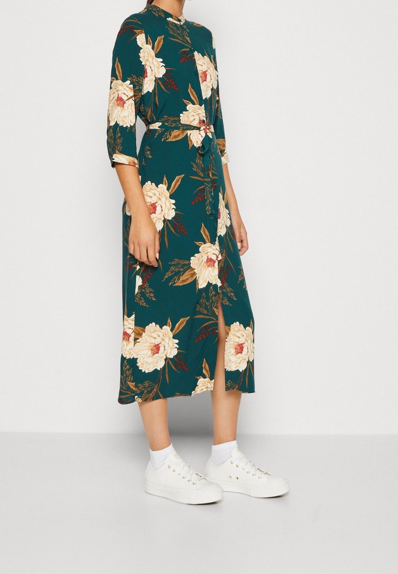 Abito midi floreale in teal con fiori crema e oro, con chiusura a bottoni davanti, vita con cintura e spacco laterale. Abbinato a scarpe da ginnastica bianche.