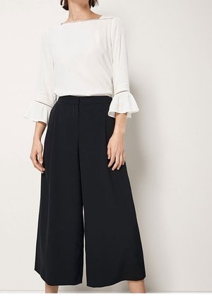 Donna che indossa una camicetta bianca con maniche a campana a tre quarti e pantaloni neri a gamba larga, a vita alta e corti, su sfondo uniforme.