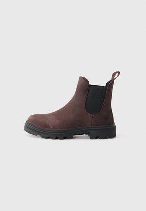 Bruine suède Chelsea-boot met zwarte elastische zijpanelen, dikke robuuste zwarte zool en een trekband aan de achterkant op een neutrale achtergrond.