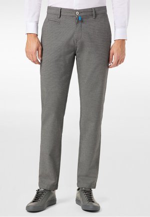 Homme portant un pantalon gris texturé, une chemise blanche et des baskets gris foncé, debout devant un fond uni.