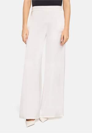 Kobieta ubrana w białe spodnie z wysokim stanem i szerokimi nogawkami, zestawione z białymi szpilkami z ostrym noskiem, ręce swobodnie opuszczone wzdłuż ciała.