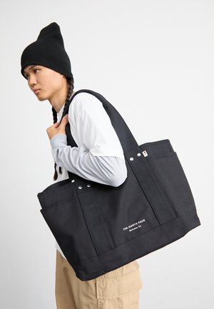 Zwarte canvas schoudertas met twee stevige handvatten, voorzien van metalen accenten en voorzakken. De merknaam is aan de onderkant in witte tekst weergegeven.
