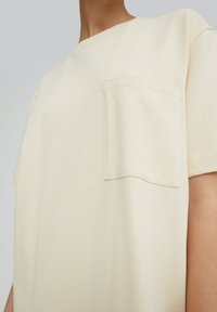Lichtbeige t-shirt van zachte katoen, met een ronde hals en een borstzak. De stof heeft een gladde textuur met minimale stikdetails.