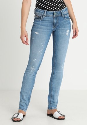 Personne portant un jean skinny bleu clair usé, un top à pois noir et blanc, et des sandales blanches à enfiler avec des boucles.