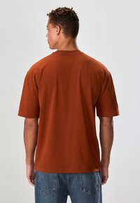 Bomullst-shirt i rostfärg med korta ärmar, rund halsringning och avslappnad passform, visad bakifrån mot en ljus bakgrund.