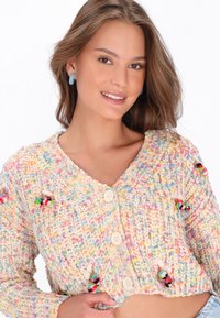 Junge Frau lächelt, trägt eine mehrfarbige Strickjacke mit Knöpfen und blaue Creolen, vor einem weißen Hintergrund.